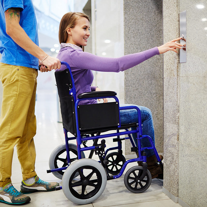 Assistant and handicapped young woman in wheelchair waiting for elevator, she pressing button by herself and smiling